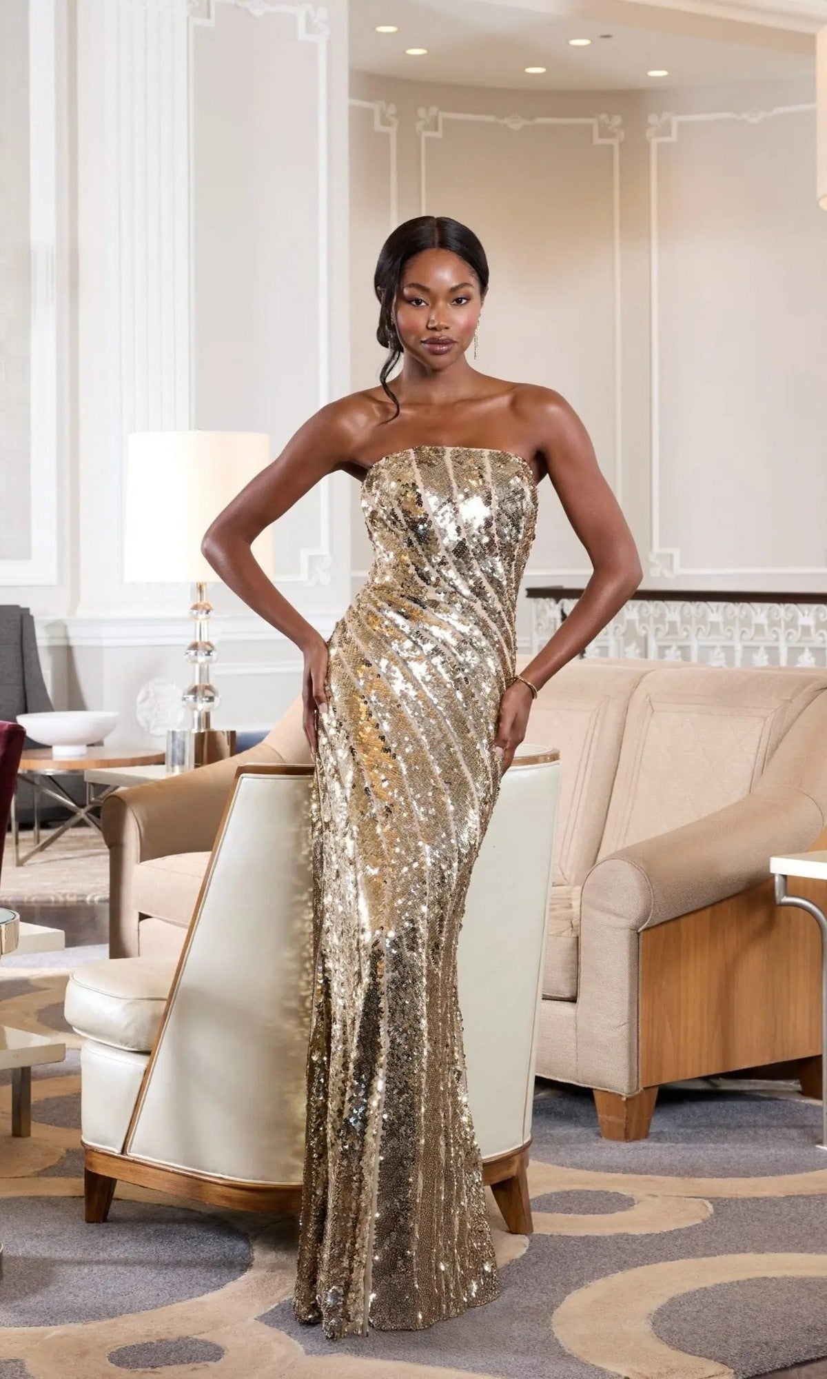 A woman stands indoors wearing the Long Formal Dress: Mac Duggal 10130, a breathtaking strapless sequin evening gown that cascades to the floor in shimmering gold. Posing with one hand on her hip in a contemporary, elegantly decorated room, she exudes the sophistication and glamour embodied by this iconic design.